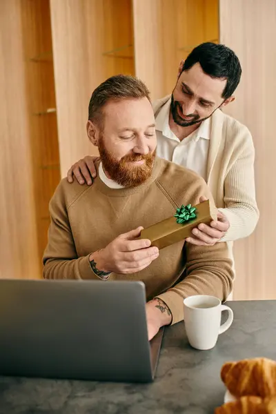 Two men exchange a gift in front of a laptop in a modern apartment, sharing a moment of happiness and love. — Stock Photo