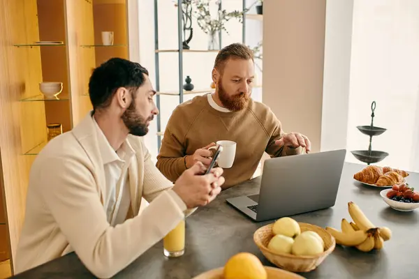 Un couple gay heureux collaborant sur un ordinateur portable à une table dans un appartement moderne. — Photo de stock