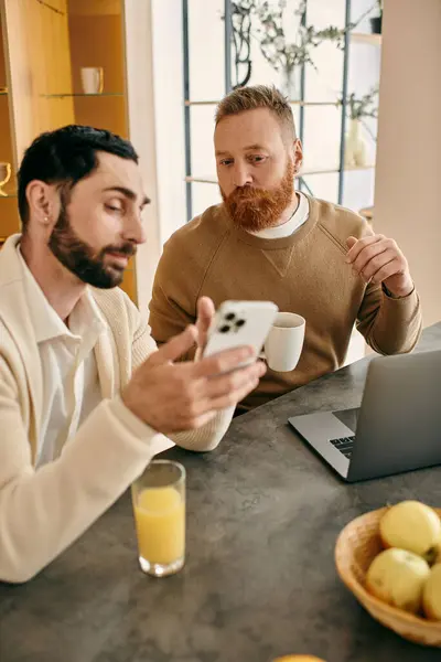 Двоє чоловіків, щаслива пара-геї, сидячи за столом в сучасній квартирі, захопилися своїми телефонами. — стокове фото