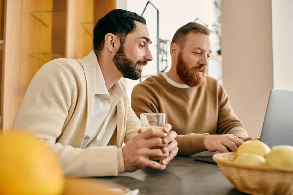A happy gay couple is seated at a table, focused on a laptop screen in a modern apartment setting. — Stock Photo
