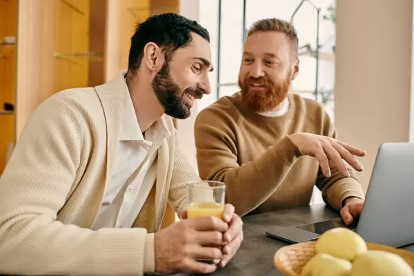 Two men happily engaged, sitting at a table, absorbed in their laptop screen in a modern apartment setting. — Stock Photo
