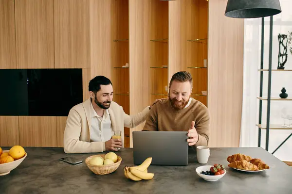 Deux hommes heureux, un couple gay, assis à une table dans un appartement moderne, absorbé par l'utilisation d'un ordinateur portable. — Photo de stock