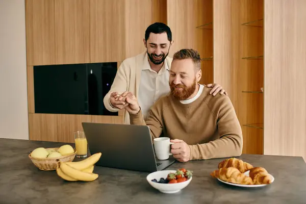 Two men, a happy gay couple, are working side by side on a laptop in their modern kitchen. — Stock Photo