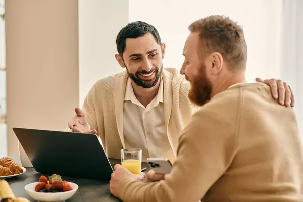 A happy gay couple sits at a table in a modern apartment, focusing on their laptop, engrossed in productive work. — Stock Photo