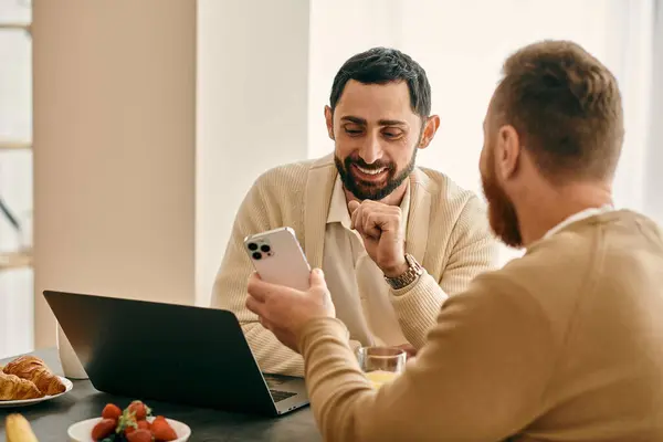 Two men, a happy gay couple, sit at a table in a modern apartment, engrossed in the screen of a laptop. — Stock Photo