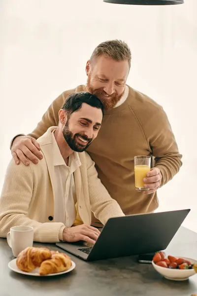 Two men, a happy gay couple, sit at a table with a laptop, engaged in work. They are also enjoying glasses of orange juice. — Stock Photo
