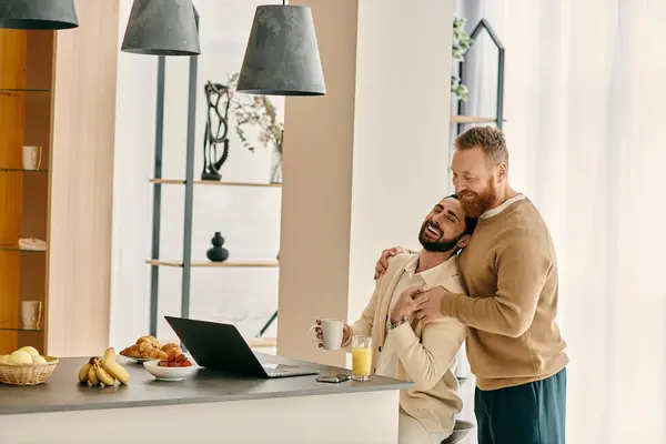 Dos hombres gay felices se abrazan mientras colaboran en una computadora portátil en una cocina moderna, mostrando amor y trabajo en equipo. - foto de stock