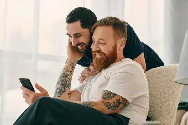 Deux hommes, un couple gay heureux, assis sur un canapé ensemble en regardant un téléphone portable dans un salon moderne. — Photo de stock