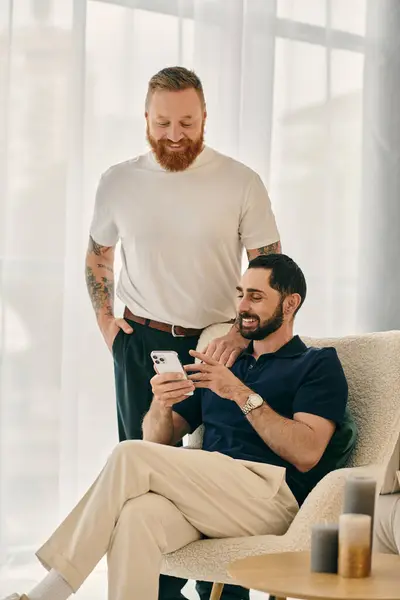 Two happy gay men in casual clothes sitting on a couch, absorbed in phone, sharing moments of love and modern connection. — Stock Photo