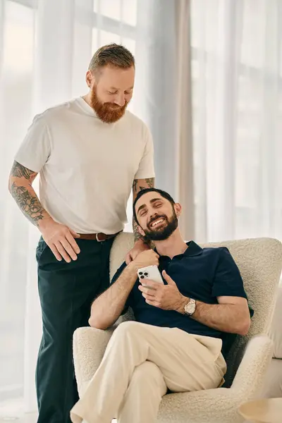 Two men, one holding a cell phone, sitting in a chair in a modern living room, sharing a happy and intimate moment. — Stock Photo