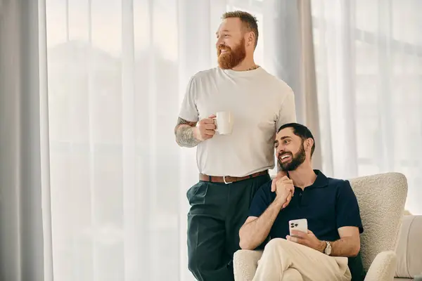 A happy gay couple in casual clothes sitting on a chair, drinking coffee, and sharing a moment in a modern living room. — Stock Photo