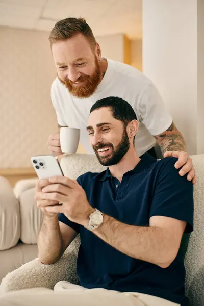 Una feliz pareja gay con ropa casual se sienta en un sofá, absorta en un teléfono celular, disfrutando de un momento de conexión moderna. - foto de stock