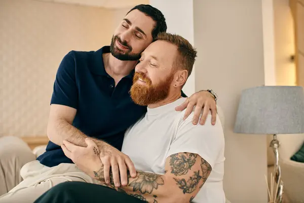 Two men with tattoos embrace warmly on a couch in a modern living room, sharing a moment of love and connection. — Stock Photo