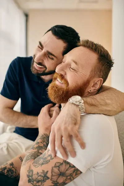 Dos hombres con tatuajes se abrazan cariñosamente en un cómodo sofá en una moderna sala de estar, reflejando el amor y la felicidad. - foto de stock