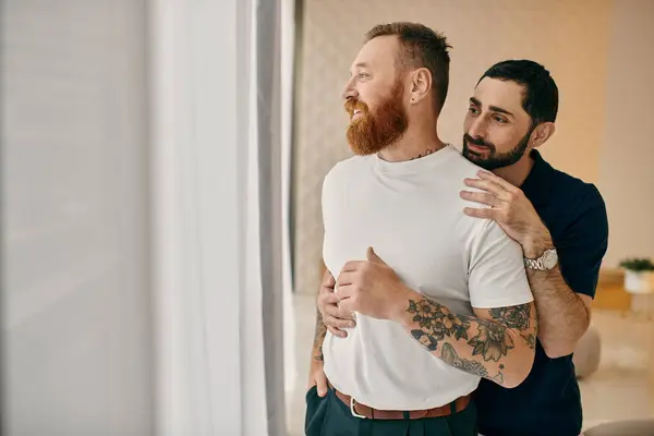 Dos hombres con ropa casual se abrazan en un cálido abrazo frente a una ventana, mostrando su amor en un moderno entorno de sala de estar. - foto de stock