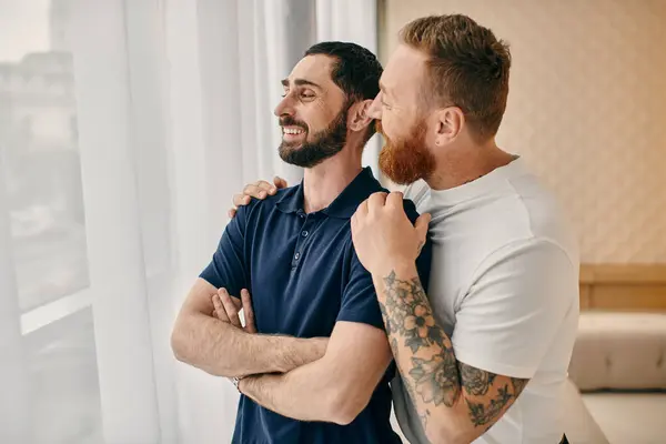 Two men, a happy gay couple, in casual clothes, embracing in front of a window in a modern living room. — Stock Photo