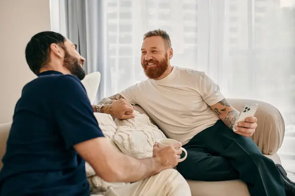 Two men in casual clothes, a happy gay couple, sit on a couch in a modern living room, engaging in a lively conversation. — Stock Photo