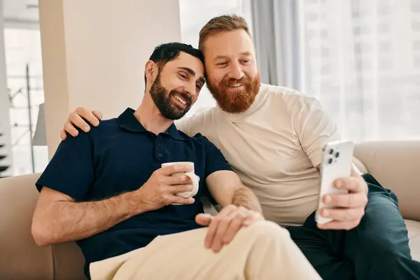 Deux hommes gays heureux en vêtements décontractés assis sur un canapé, absorbé dans leur téléphone portable, partageant un moment de connexion moderne. — Photo de stock