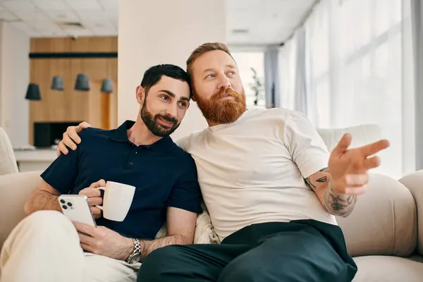 Happy gay couple in casual clothes, sitting on a couch with a cup of coffee in a modern living room. — Stock Photo
