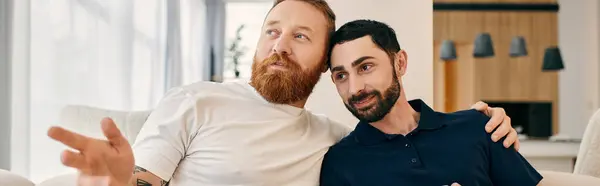 Two bearded men in casual attire sit closely on a couch in a modern living room, enjoying quality time together. — Stock Photo