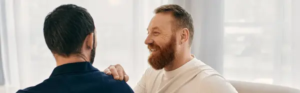 Two men with beards, one in a gray shirt and the other in a black shirt, joyfully smiling at each other in a cozy living room setting. — Stock Photo