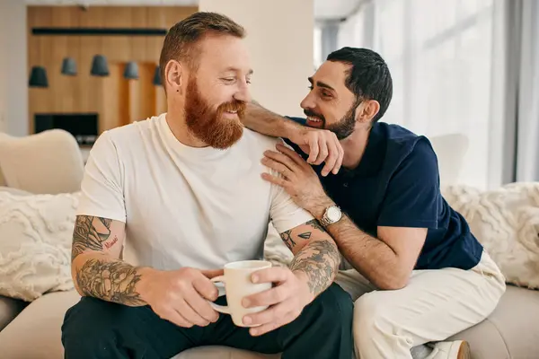 A gay couple in casual attire relax on a couch, enjoying each others company and sipping coffee in a modern living room setting. — Stock Photo