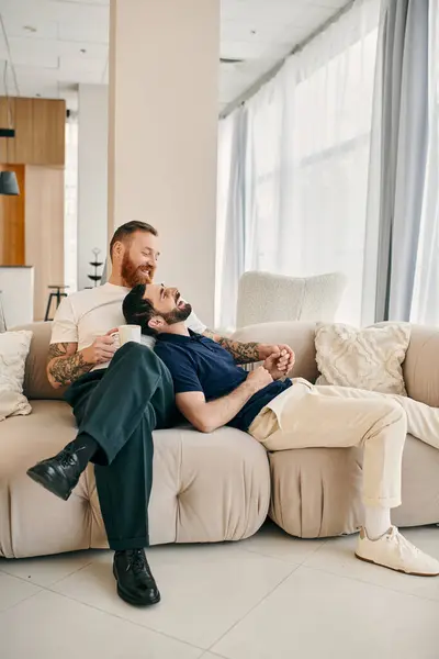 Two men, a happy gay couple, dressed casually, sit together on a modern living room couch, enjoying each others company. — Stock Photo