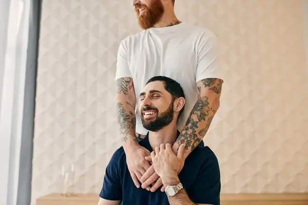 A man with tattoos and a beard is sitting on another mans shoulder, both smiling in their casual attire in a modern living room. — Stock Photo