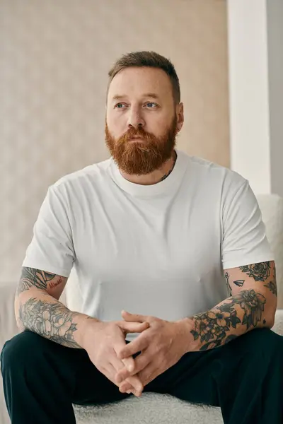 Homem barbudo com tatuagens senta-se confortavelmente em um sofá, exalando confiança e auto-expressão. — Fotografia de Stock