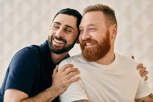 Due uomini barbuti, felici e innamorati, si abbracciano saldamente in un salotto moderno. — Foto stock