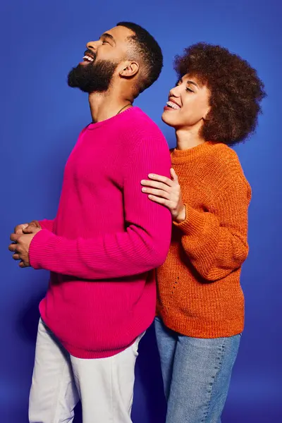 Un jeune homme et une jeune femme afro-américaine, vêtus de vêtements décontractés vibrants, partagent un moment de joie et de rire sur un fond bleu. — Photo de stock