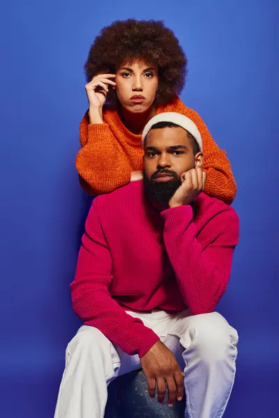 A young African American man and woman dressed in vibrant casual attire, sitting atop each other in a friendly embrace on a blue background. — Stock Photo