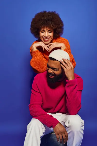 Eine junge Afroamerikanerin in lebendiger Kleidung sitzt auf den Schultern eines Mannes, beide lächeln in einem Moment der Freundschaft. Blauer Hintergrund. — Stockfoto