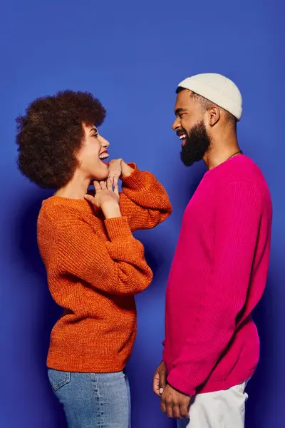 A young African American man and woman in vibrant casual attire sharing a moment of laughter in front of a blue background. — Stock Photo