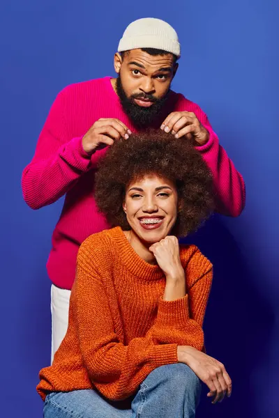 Ein junger Afroamerikaner und eine junge Afroamerikanerin sitzen zusammen und präsentieren lebhafte Freizeitkleidung und ein starkes Freundschaftsband auf blauem Hintergrund.. — Stockfoto