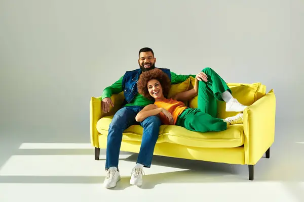 Un homme et une femme afro-américains souriants, vêtus de vêtements colorés, assis ensemble sur un canapé jaune sur un fond gris. — Photo de stock