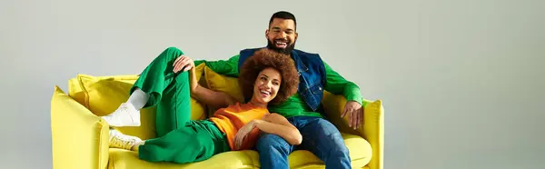 Happy amici afroamericani in abiti vibranti seduti su un divano giallo su uno sfondo grigio. — Foto stock