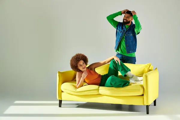 A man and woman, dressed in vibrant clothes, share a moment of friendship while standing and sitting on a yellow couch. — Stock Photo