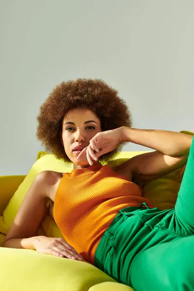 Una mujer afroamericana rizada se sienta en la parte superior de un sofá amarillo brillante, mostrando relajación. - foto de stock