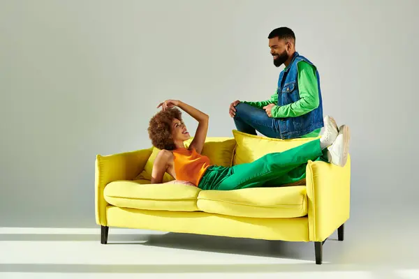 A happy African American man and woman in vibrant clothes sit on a yellow couch against a grey background. — Stock Photo