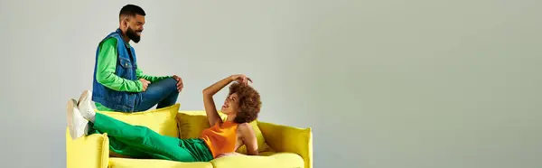 Happy African American friends in vibrant clothes sitting on a yellow couch against a grey background. — Stock Photo