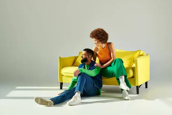Un homme et une femme afro-américains heureux en vêtements vibrants assis ensemble sur un canapé jaune sur un fond gris. — Photo de stock