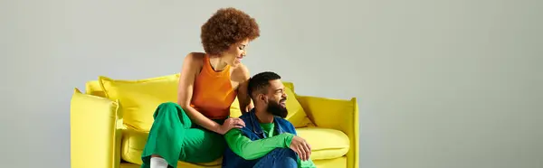 Heureux amis afro-américains en vêtements vibrants assis sur une chaise jaune sur un fond gris. — Photo de stock