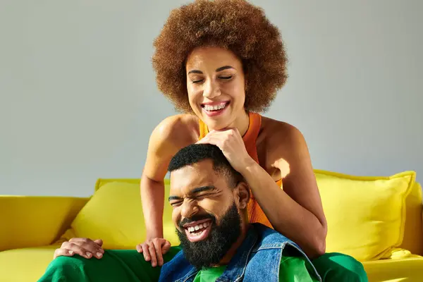 A man and a woman, both African American, laughing and chilling on a bright yellow couch against a grey backdrop. — Stock Photo
