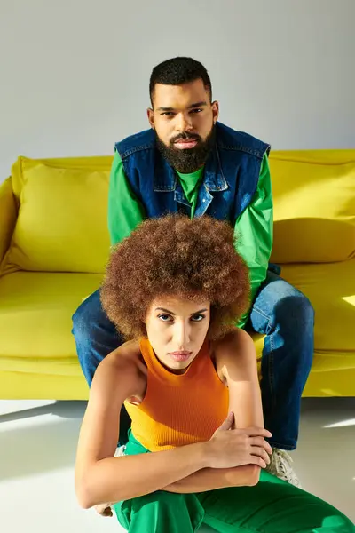 Afro-Américain homme et femme en tenue colorée assis ensemble sur un canapé jaune sur un fond gris. — Photo de stock
