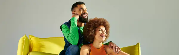 Afroamerikanische Freunde in bunten Kleidern teilen einen Moment der Freundschaft auf einer gelben Couch vor grauem Hintergrund. — Stockfoto