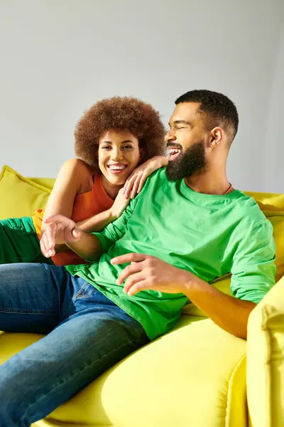 Un uomo e una donna afroamericani, vestiti vivacemente, condividono un momento di amicizia seduti su un divano giallo su uno sfondo grigio. — Foto stock