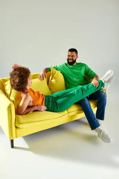 Радостные афроамериканец мужчина и женщина в яркой одежде разделяют момент дружбы на желтом диване на сером фоне. — стоковое фото