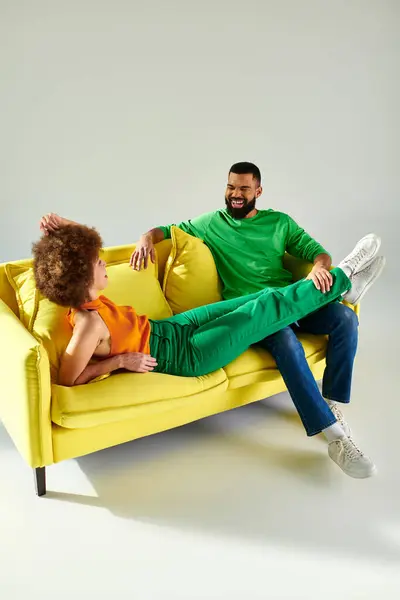 A joyful African American man and woman in vibrant clothes share a moment of friendship on a yellow couch against a grey background. — Stock Photo