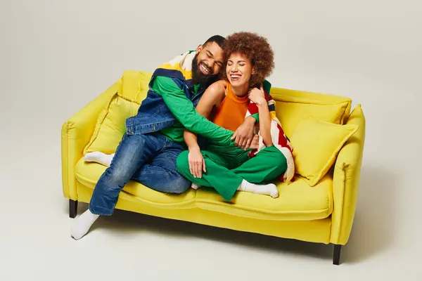 A man and woman, dressed in colorful attire, sit happily together on a yellow couch against a grey background. — Stock Photo
